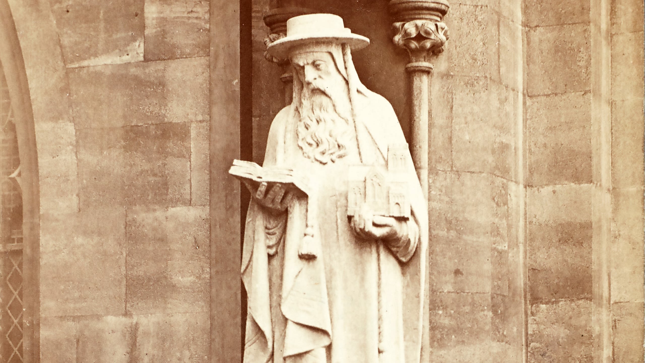 A stone statue of St. Peter holding a book and a model of a cathedral building, set against a stone wall with decorative columns.